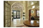Spacious and elegant interior entryway of a senior living facility with a large arched glass door, stone wall accents, tiled floor, potted plants, and a round wooden table in the foreground. A living area with seating and curtains is visible to the right.