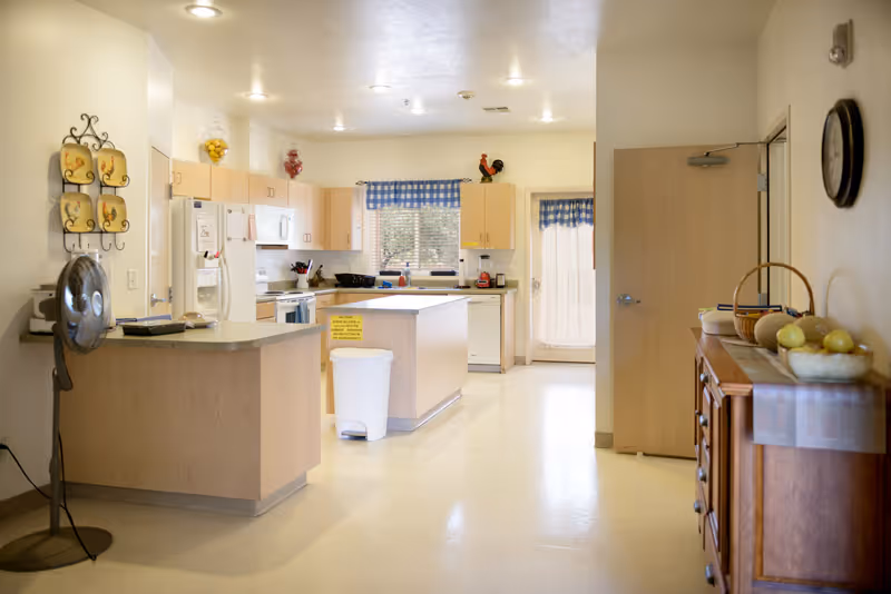 Bright and clean kitchen area in an assisted living facility with light wood cabinets, a white refrigerator, stove, dishwasher, and a central island. The room has a window with blue checkered curtains and a door with a sheer curtain. A standing fan is positioned near the counter, and a wooden sideboard with a basket of fruit and a clock on the wall is visible on the right.