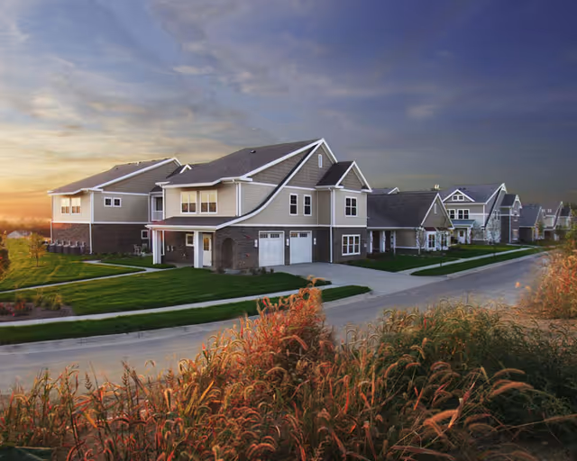 Row of two-story residential buildings with garages and landscaped lawns under a colorful sunset sky.