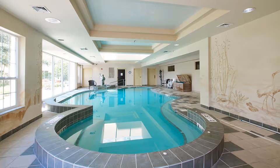 Bright indoor curved swimming pool with tiled decking, large windows, seating, and a wall mural.