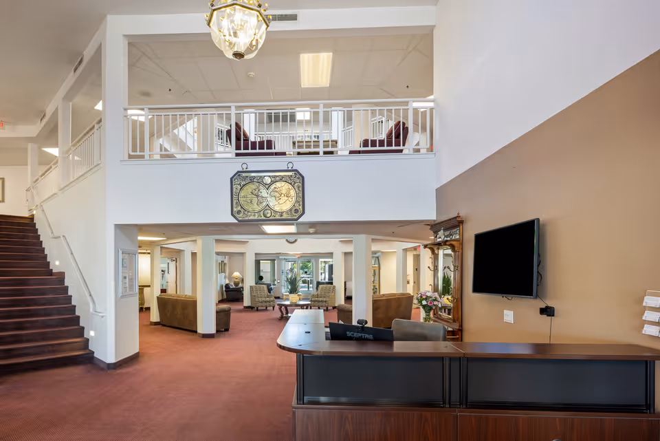 Interior view of a senior living community lobby with a reception desk in the foreground, a mounted flat-screen TV on the right wall, seating areas with sofas and armchairs in the background, a staircase on the left, and a balcony with additional seating above.