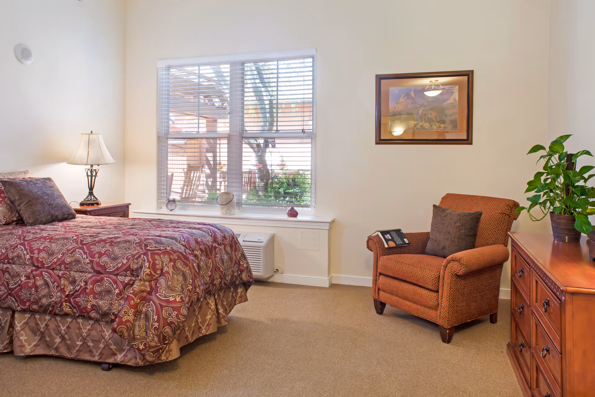 A cozy bedroom with a patterned bed, bedside lamp, armchair, window with blinds, and a wooden dresser with a plant.