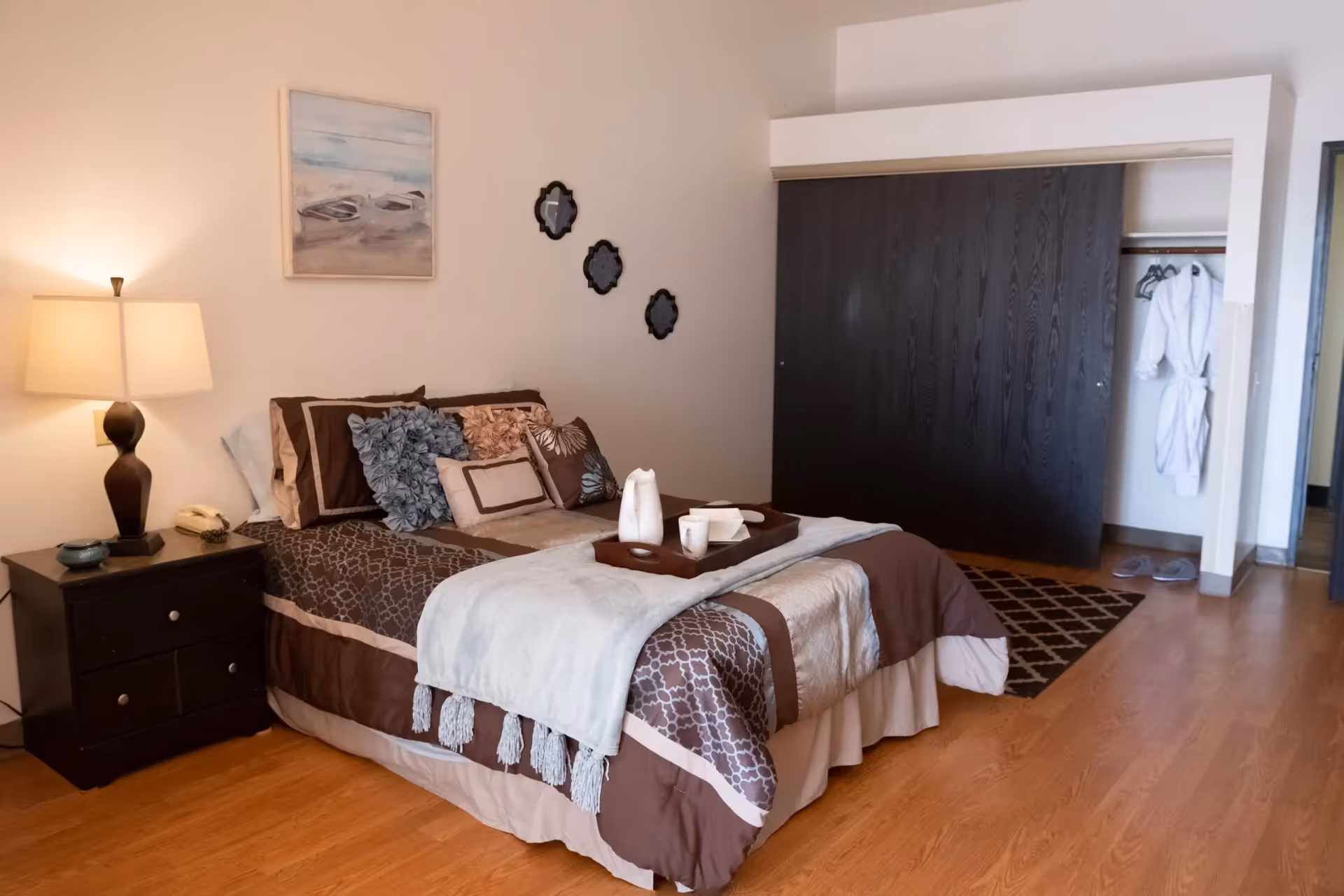 A cozy bedroom with a neatly made bed featuring brown and beige bedding and multiple decorative pillows. A wooden nightstand with a lamp and a telephone is beside the bed. On the wall above the bed are three small decorative mirrors and a framed painting of boats on water. There is a closet with dark sliding doors partially open, revealing white robes and slippers inside. The room has wooden flooring and a patterned rug near the closet.