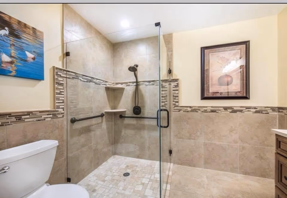A modern bathroom featuring a walk-in shower with glass door and tiled walls and floor. The shower has a black showerhead and two black grab bars for support. There is a white toilet to the left and a wooden vanity with a countertop to the right. The walls are decorated with a framed picture and a painting of ducks on water.