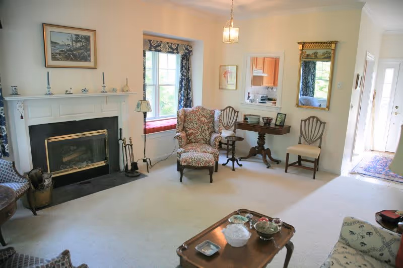 A cozy living room with a fireplace on the left wall, a window with a cushioned bench seat, an upholstered armchair with a matching footrest, two wooden chairs, a small table with framed photos, and a coffee table with decorative items. The room is carpeted and has light-colored walls with a mirror and framed artwork.