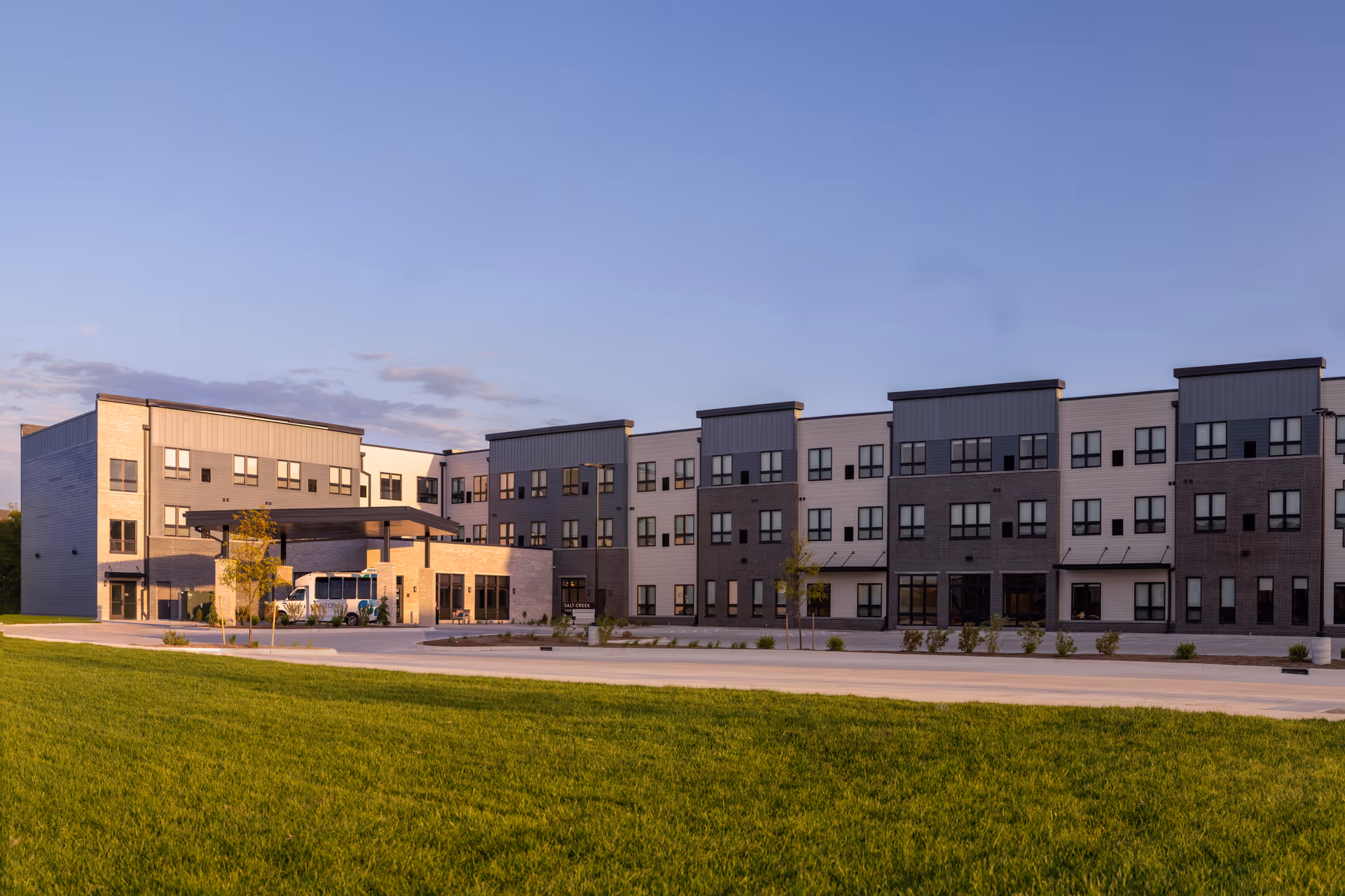 Exterior view of a modern three-story senior living facility building named Salt Creek Senior Living, with a covered entrance and a shuttle bus parked outside, surrounded by a well-maintained lawn and young trees under a clear sky at sunset.