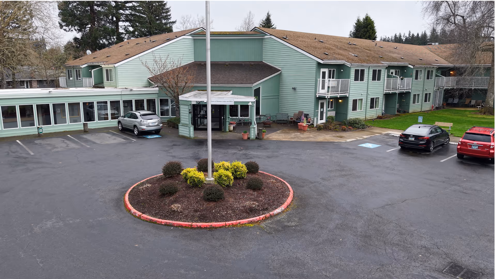 Front exterior of a mint-green two-story senior living building with a circular planter, flagpole and parked cars in the lot.
