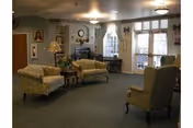 Communal living room with upholstered chairs and sofas arranged around a coffee table, a fireplace, wall clock, lamps, and large windows.