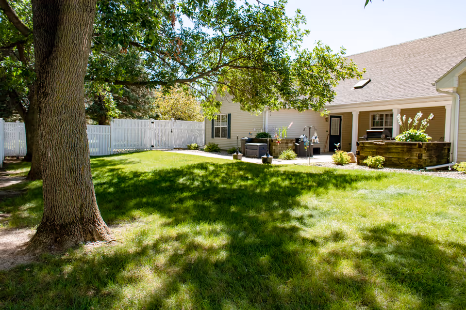 A sunny outdoor garden area with green grass, large trees providing shade, and a beige building with a covered patio. The patio has potted plants, a grill, and outdoor furniture. A white fence encloses the yard.