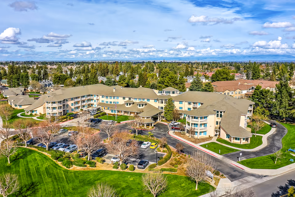 Aerial view of Solstice Senior Living at Bakersfield, showing a large, multi-story senior living facility surrounded by green lawns, trees, and a parking lot with several cars. The building has a beige exterior with a gray roof and is situated in a suburban area with other houses and trees in the background under a partly cloudy sky.