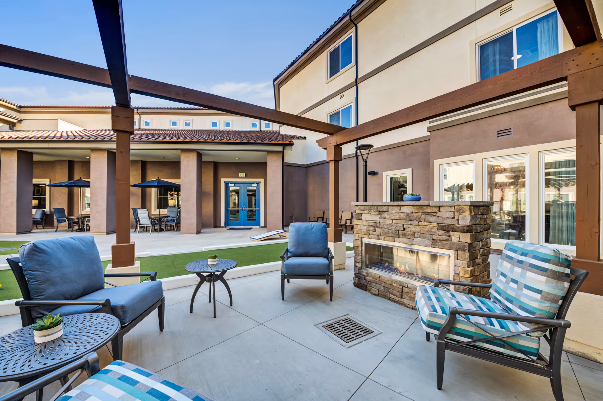 Outdoor courtyard seating area with cushioned chairs, a stone fireplace, pergola beams, and the adjacent building facade.