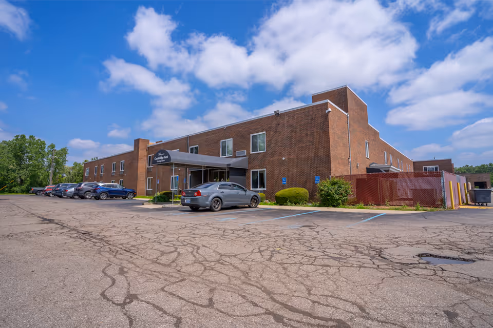Exterior view of a two-story brick building under a partly cloudy blue sky. Several cars are parked in front of the building, which has a covered entrance with a sign that reads 'Mission Continuing Care Nursing Home.' The parking lot shows visible cracks and patches.