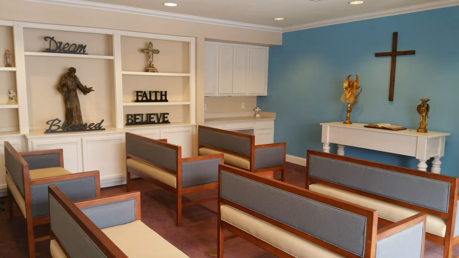 A small chapel room with wooden pews arranged in rows facing a white altar table against a blue wall. A wooden cross hangs on the wall above the altar, which holds an open book and two golden decorative items. On the left side, white built-in shelves display religious statues and words like 'Dream', 'Faith', 'Believe', and 'Blessed'.