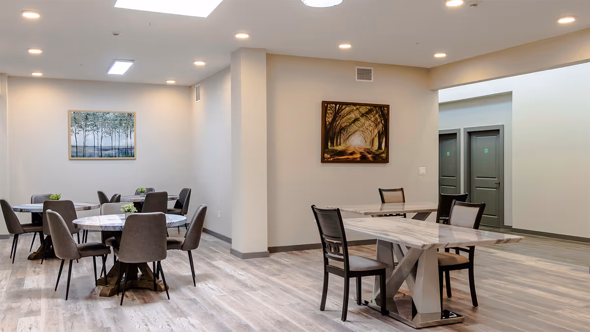 A clean and modern dining area with multiple tables and chairs arranged neatly. The room has light-colored walls, wood-style flooring, and two framed landscape paintings on the walls. There are recessed lights in the ceiling providing bright illumination.