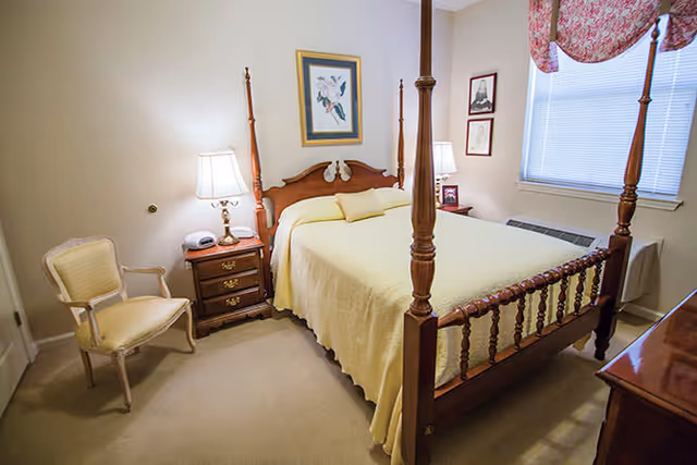A cozy bedroom with a wooden four-poster bed covered with a light yellow bedspread. There are two bedside tables with lamps on each side of the bed. A framed picture hangs above the headboard. A cushioned armchair is placed near the left wall, and a window with blinds and a floral valance is on the right wall.