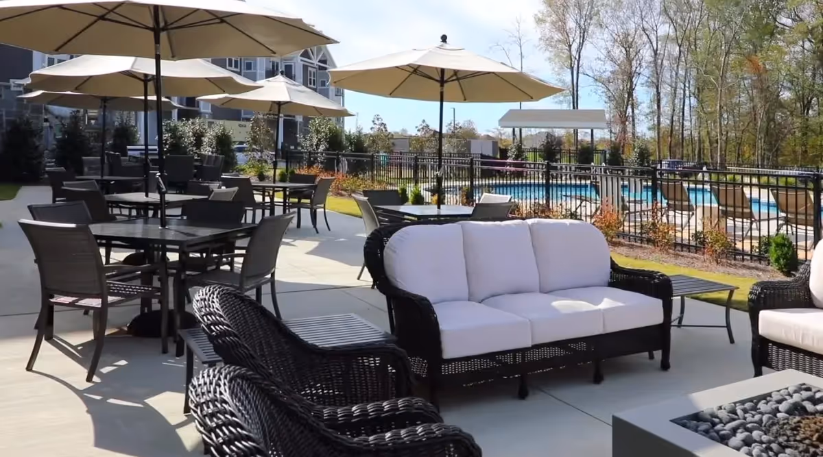 Outdoor patio area with multiple tables and chairs under large umbrellas, cushioned wicker seating, a fire pit, and a fenced swimming pool in the background surrounded by trees and landscaping.