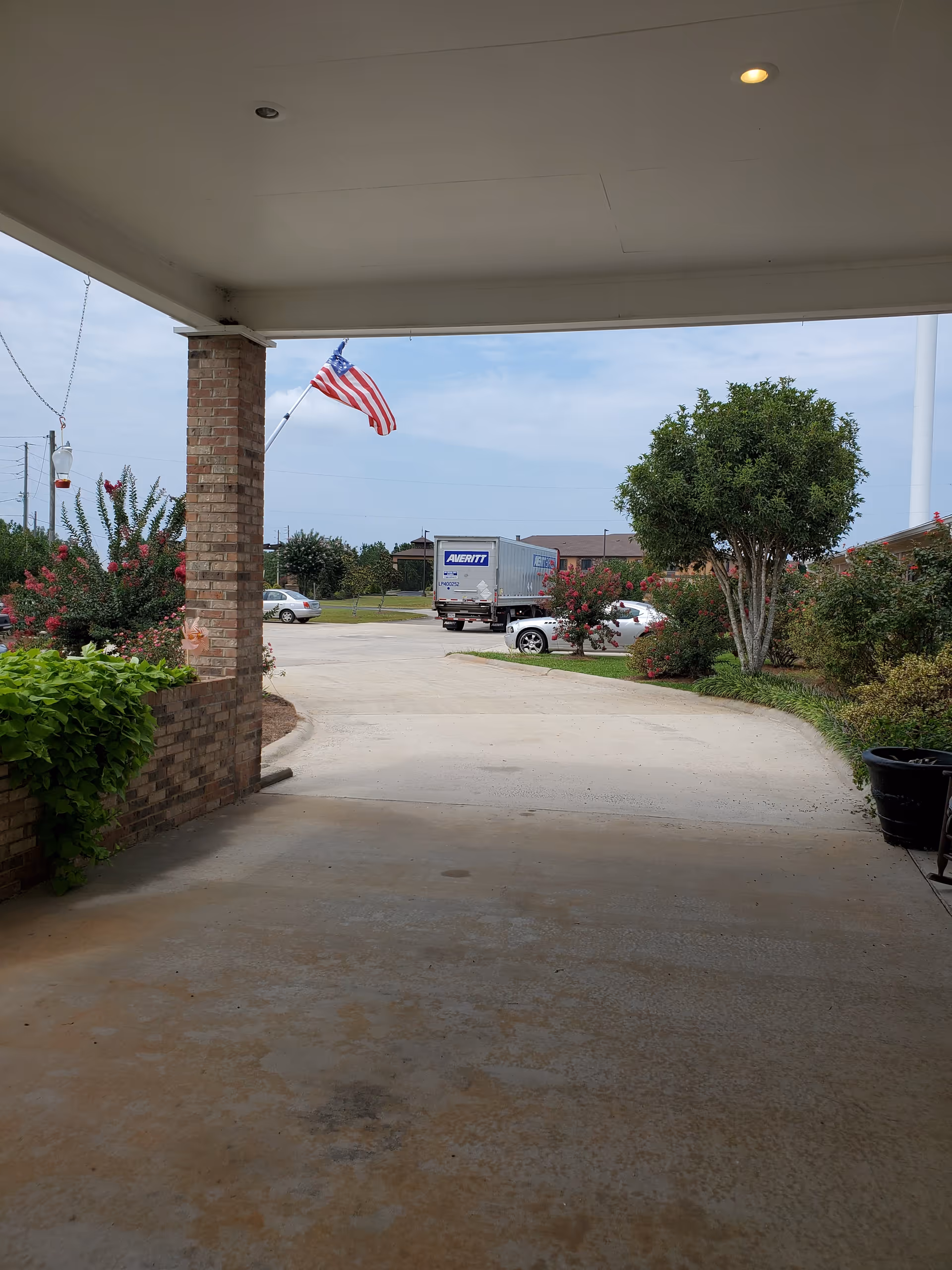 View from under a covered driveway or porch area looking out towards a parking lot with a truck and cars. There is a brick column on the left, green bushes and flowering plants on both sides, and an American flag flying on a pole. The sky is partly cloudy.