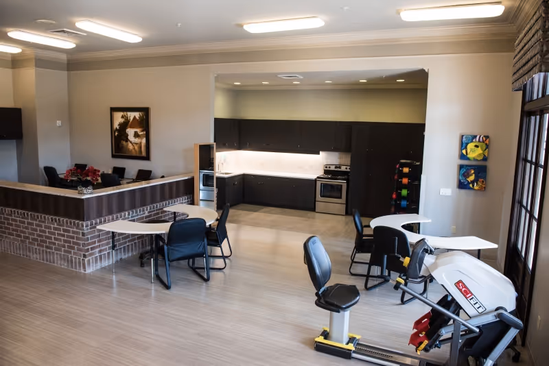 Interior view of a senior living facility common area with a small kitchen in the background featuring dark cabinets and stainless steel appliances. In the foreground, there are tables with chairs and an exercise machine near large windows with blinds. The room has light-colored flooring and neutral walls with some artwork.