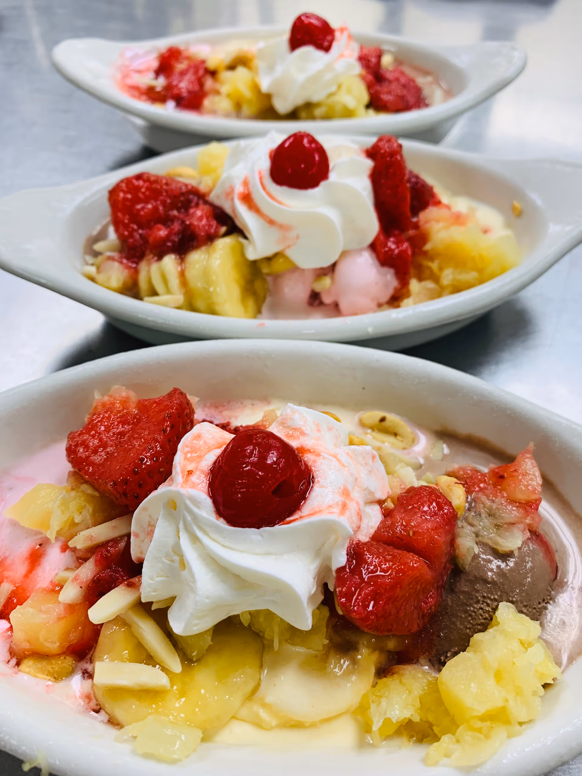 Three banana-split style sundaes topped with whipped cream, cherries, strawberries, banana slices and pineapple in white dishes on a metal surface.