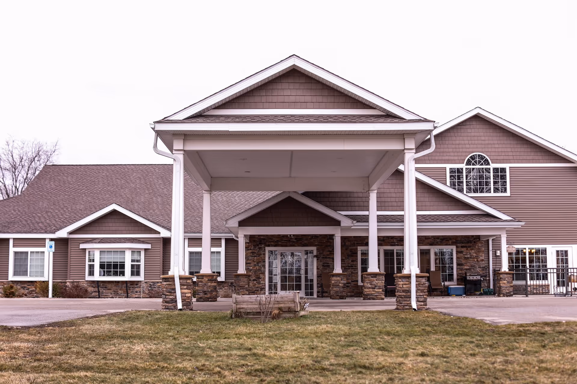 Front exterior view of Reed City Fields facility showing a large covered entrance with white pillars and stone bases, brown siding, multiple windows, and a grassy area in front.