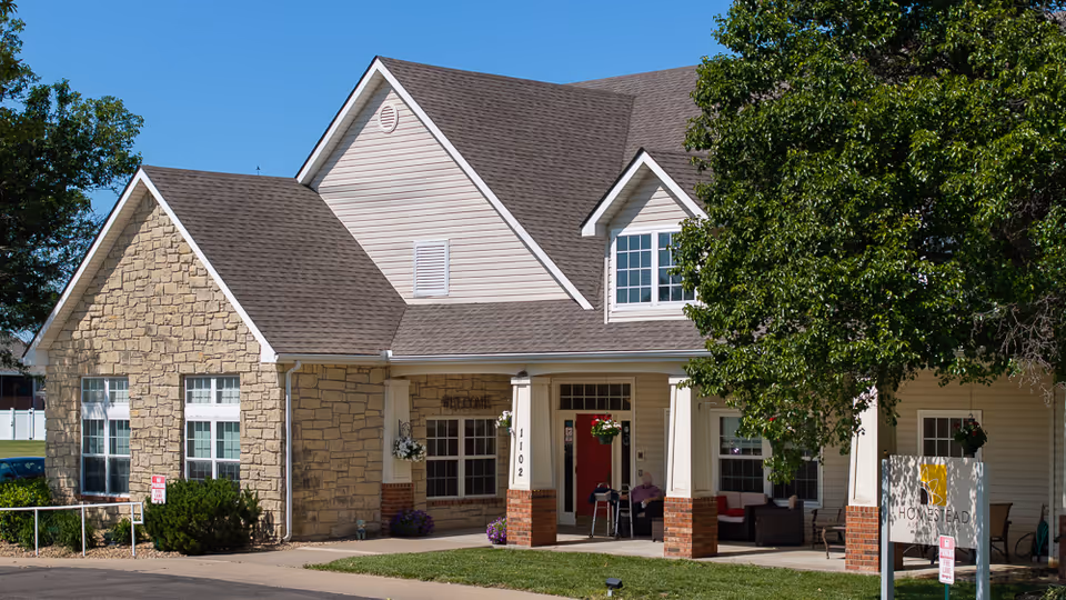 Exterior view of Homestead Assisted Living of Abilene showing a beige building with stone and siding facade, a red front door, a covered porch with seating, and a large tree on the right side under a clear blue sky.