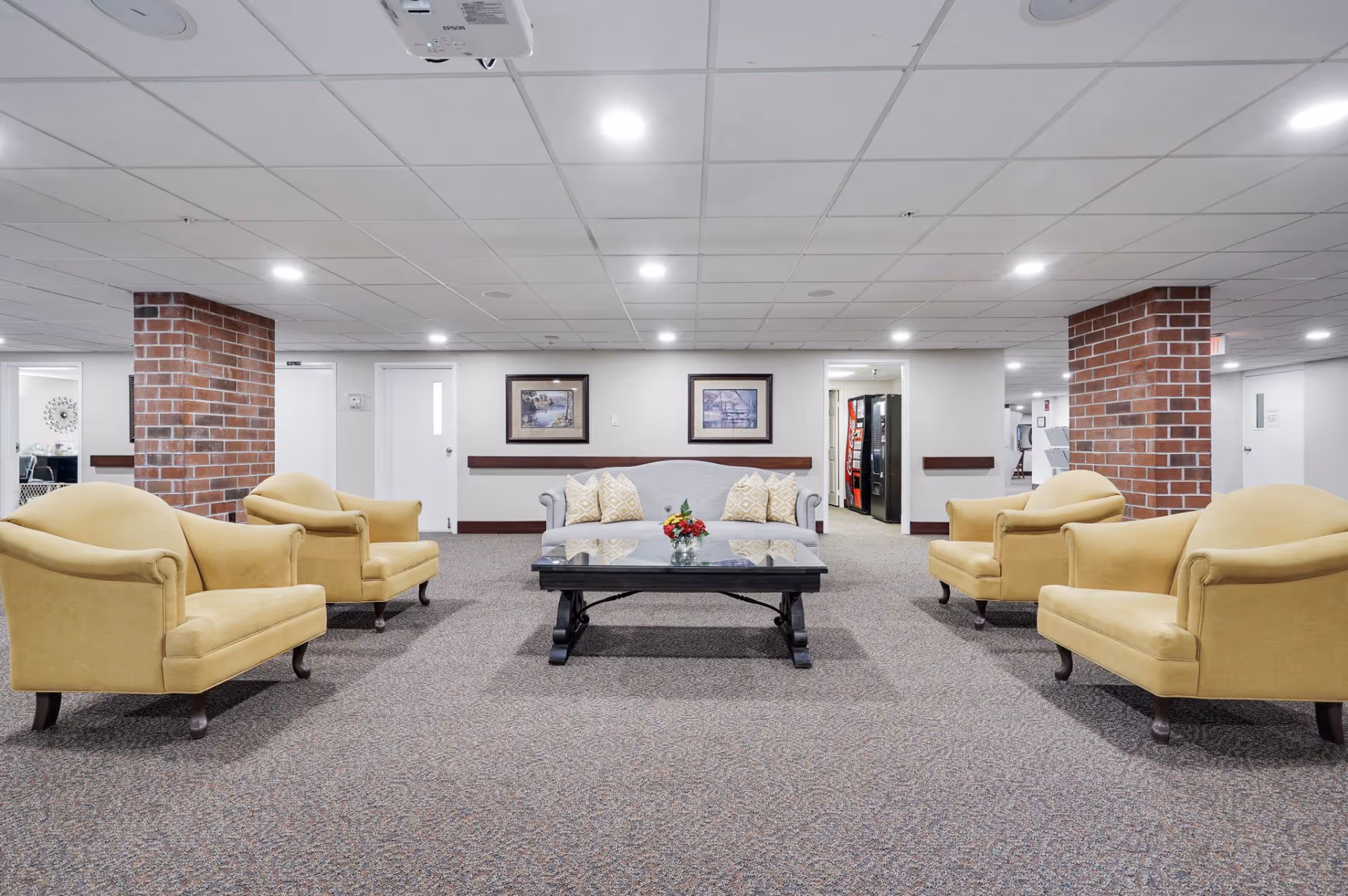 A spacious lounge area in Brownsboro Park Retirement Community featuring a gray sofa with patterned cushions, four yellow armchairs arranged around a black coffee table with a small flower arrangement. The room has carpeted flooring, white walls, two exposed brick columns, and a drop ceiling with recessed lighting. Two framed pictures hang on the wall behind the sofa, and vending machines are visible through an open doorway.