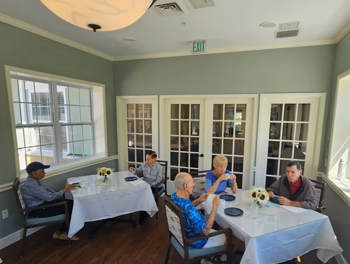 A dining room in a senior living facility with five elderly people seated around two tables covered with white tablecloths. The room has light green walls, large windows letting in natural light, and French doors in the background. There are small flower arrangements on the tables.
