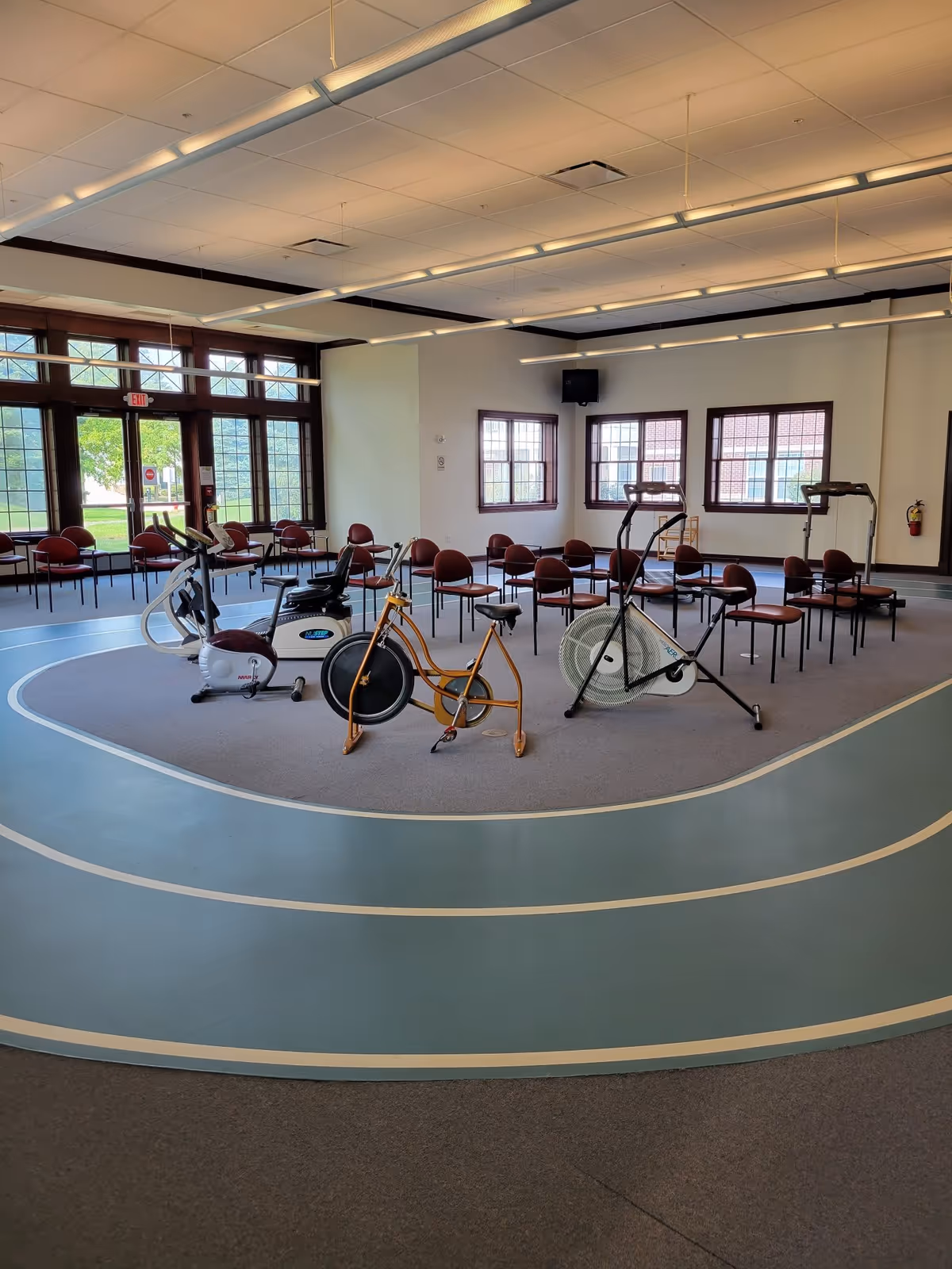 Bright multipurpose activity room with stationary exercise bikes on a small indoor walking track and rows of chairs by large windows.