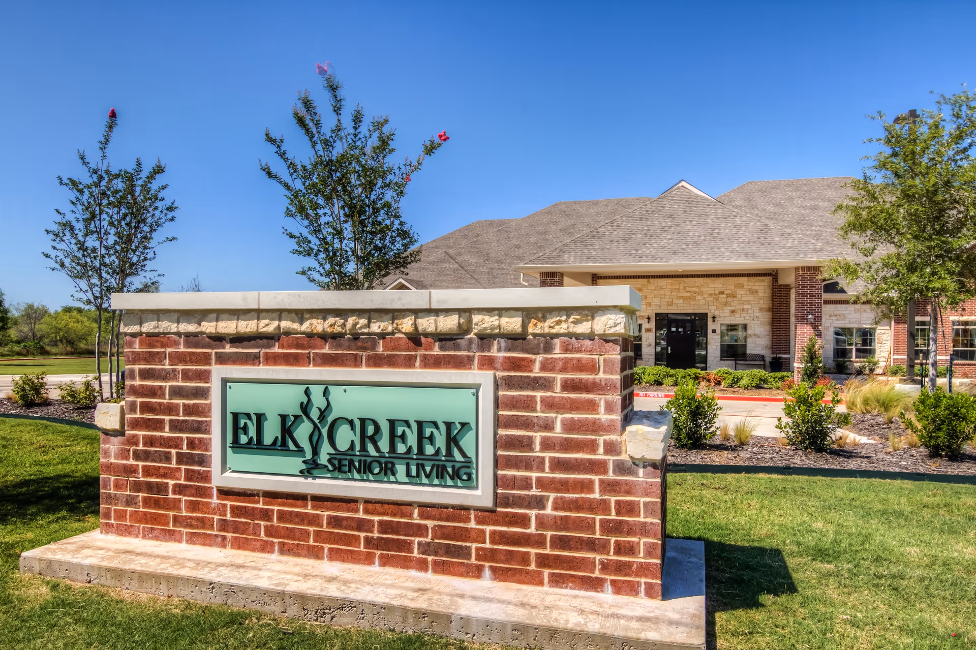 A brick sign with a stone top displaying the text 'Elk Creek Senior Living' in front of a single-story building with a gray shingled roof, beige and red brick exterior, surrounded by green grass, small bushes, and trees under a clear blue sky.