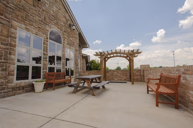 Outdoor patio area at The Pavilion Senior Living at Lebanon featuring stone exterior walls, large windows, two wooden benches, a wooden picnic table, a potted plant, and a wooden pergola under a partly cloudy sky.