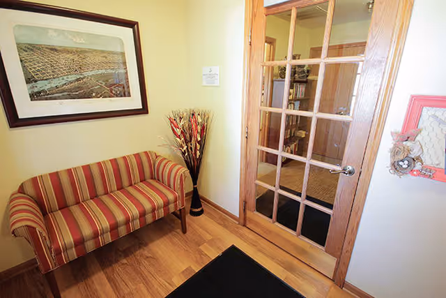 Small waiting area with a striped red and beige loveseat, a framed aerial photograph on the wall, a tall vase with decorative dried plants, and a wooden door with glass panes leading to another room with bookshelves.
