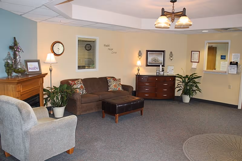 A cozy living room area in an assisted living facility featuring a brown sofa with patterned cushions, a gray armchair, a dark brown ottoman, and a wooden fireplace mantel with decorative items. There are two potted plants, a wall clock, framed artwork, and soft lighting from lamps and wall sconces. The walls are painted beige and there is a carpeted floor with a circular rug near the entrance window.