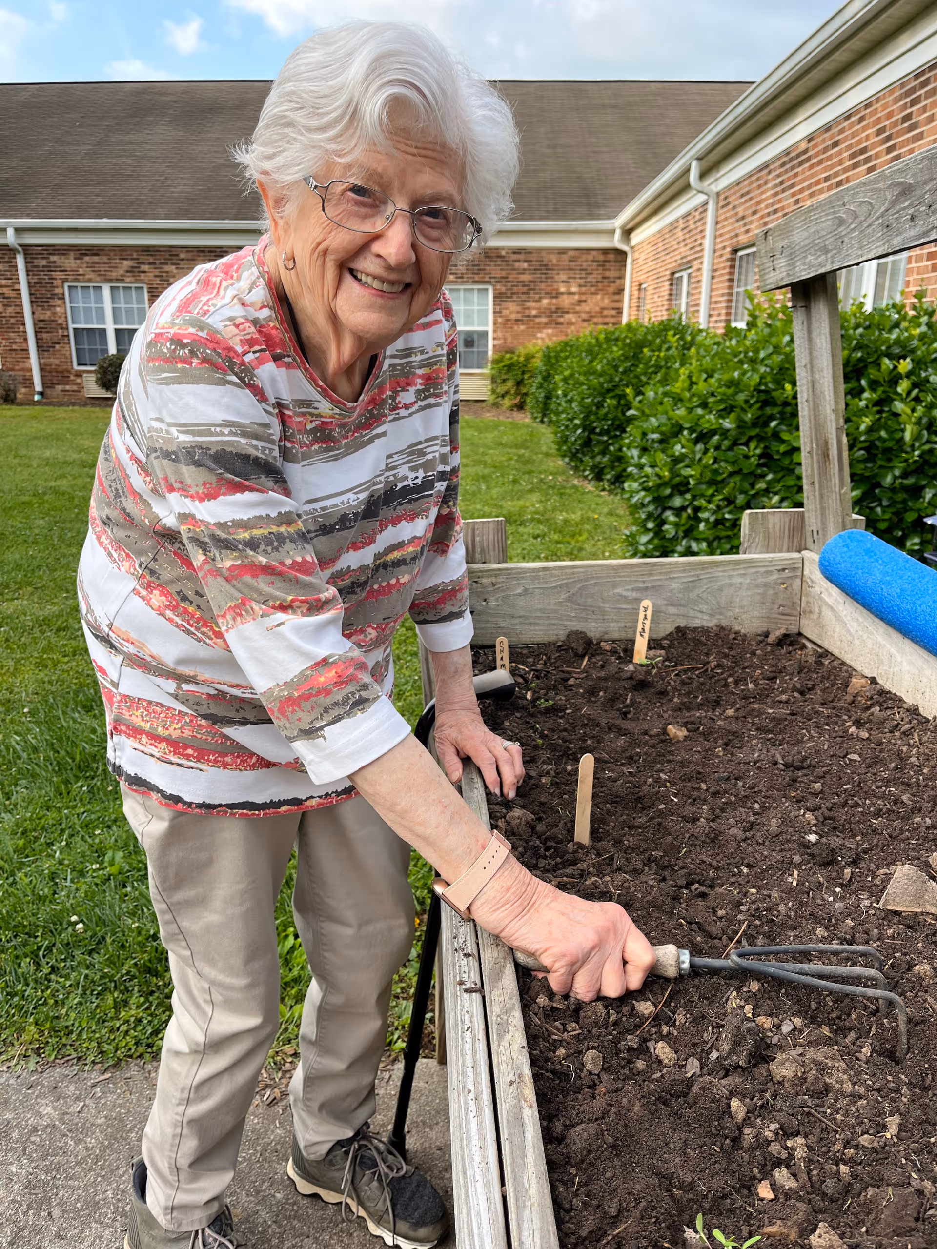 An elderly woman with white hair and glasses is smiling while gardening outdoors. She is wearing a striped long-sleeve shirt and beige pants, and is using a small gardening tool to tend to soil in a raised garden bed. Behind her is a brick building with windows and green bushes.