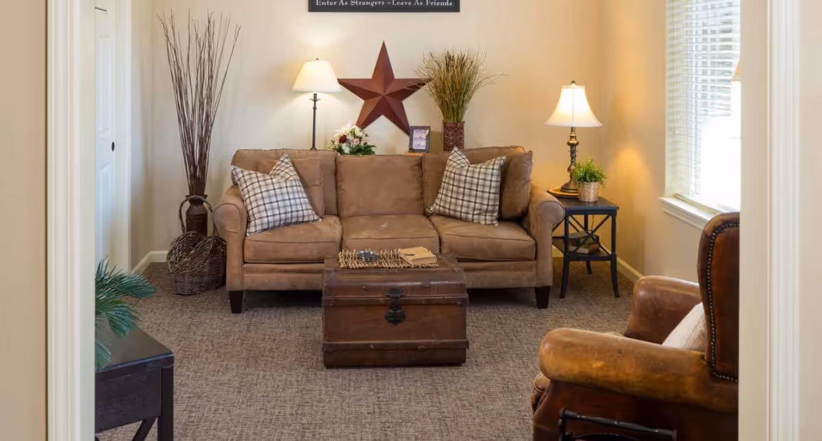 A cozy living room with a brown sofa adorned with two plaid pillows, a wooden chest used as a coffee table, a brown leather armchair, two side tables with lamps, decorative plants, and a large red star wall decoration.