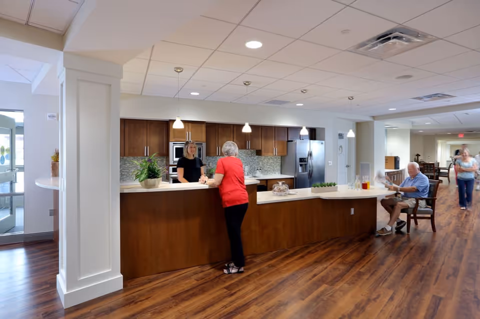 A modern kitchen area in a senior living facility with wooden cabinets, stainless steel appliances, and a long counter. An elderly woman in a red shirt is talking to a staff member behind the counter, while an elderly man sits nearby reading a newspaper. Another person is walking in the background down a hallway with wooden flooring.