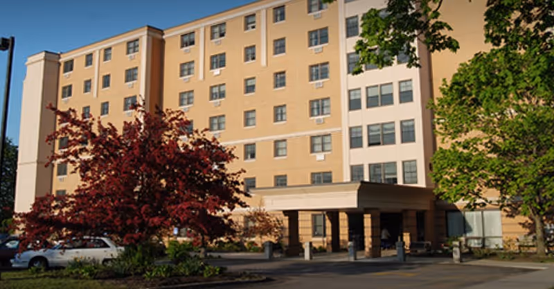 Exterior view of a multi-story senior living facility building with beige and white walls, several windows, a covered entrance, and trees with green and red foliage in front.