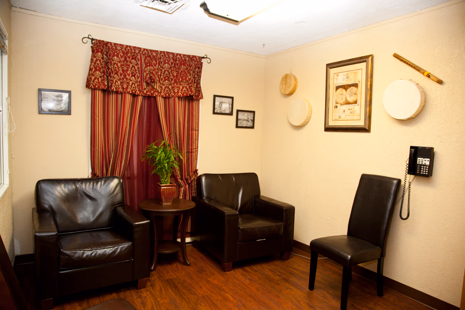 A small sitting area with two black leather armchairs and a black leather chair arranged around a round wooden side table with a potted plant. The walls are beige with framed pictures and decorative drums hanging. A red and gold patterned curtain covers the window, and a wall-mounted telephone is visible.