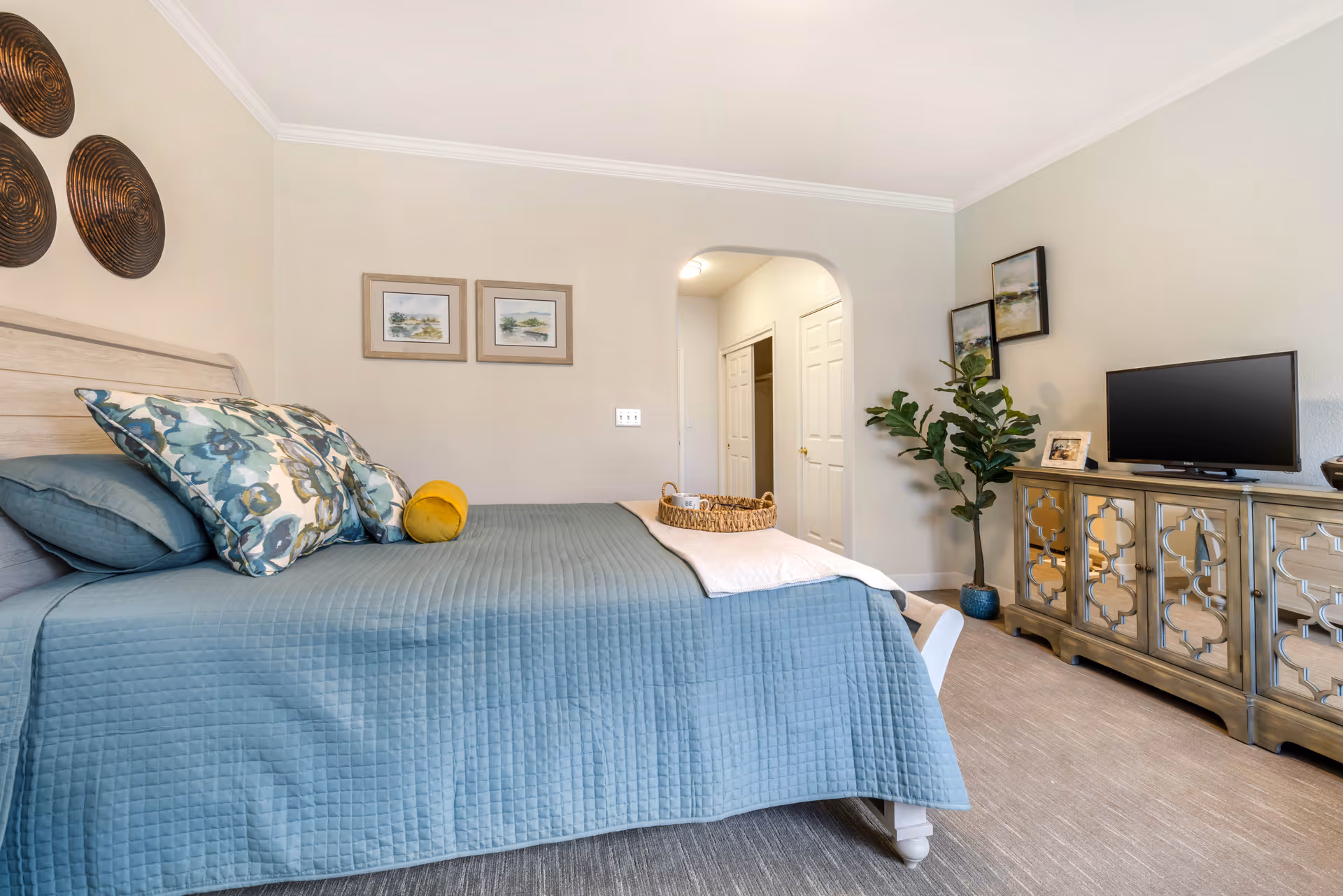 A cozy bedroom with a neatly made bed covered in a blue quilt and floral pillows. The room features light-colored walls adorned with decorative wall art, a wooden dresser with a flat-screen TV on top, a potted plant, and an arched doorway leading to a closet area.
