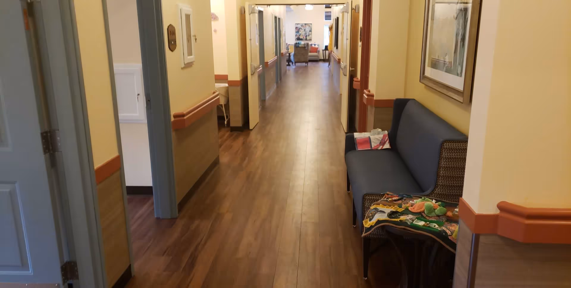 A long hallway in a senior living facility with wooden flooring and yellow walls. There are several open doors along the hallway, a blue cushioned bench with a colorful quilt and stuffed toys on the right side, and a seating area with chairs and a table visible at the far end.