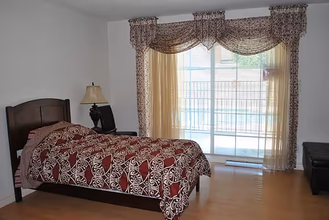A bedroom with a single bed covered in a red and white patterned bedspread, a wooden headboard, a bedside table with a lamp, and a large window with sheer and patterned curtains letting in natural light.
