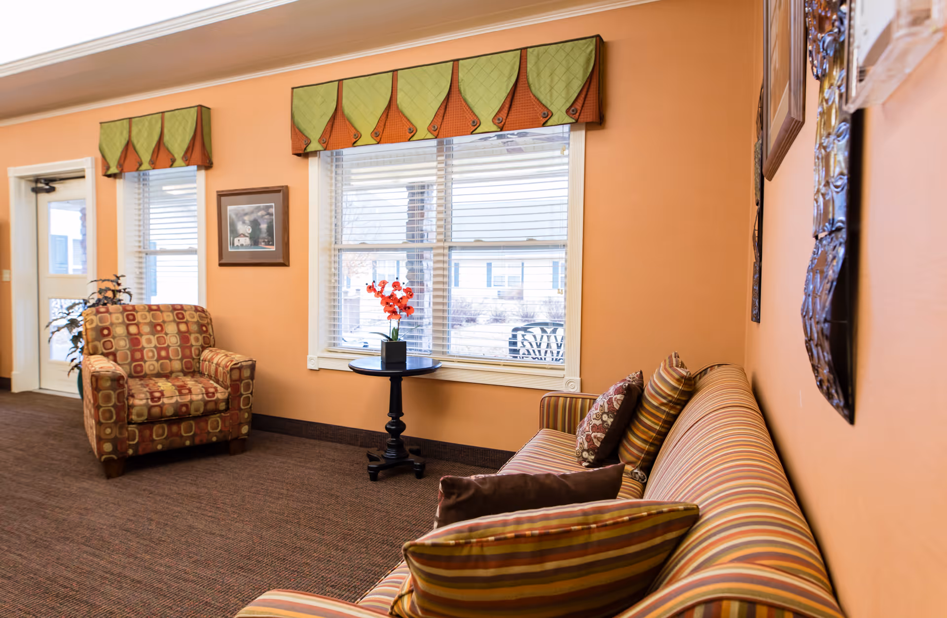 A cozy sitting area in a senior living facility with a striped sofa adorned with multiple pillows, a patterned armchair, a small round table with a vase of red flowers, and two windows with green and orange valances. The walls are painted a warm orange color and there is a door and a framed picture on the wall.