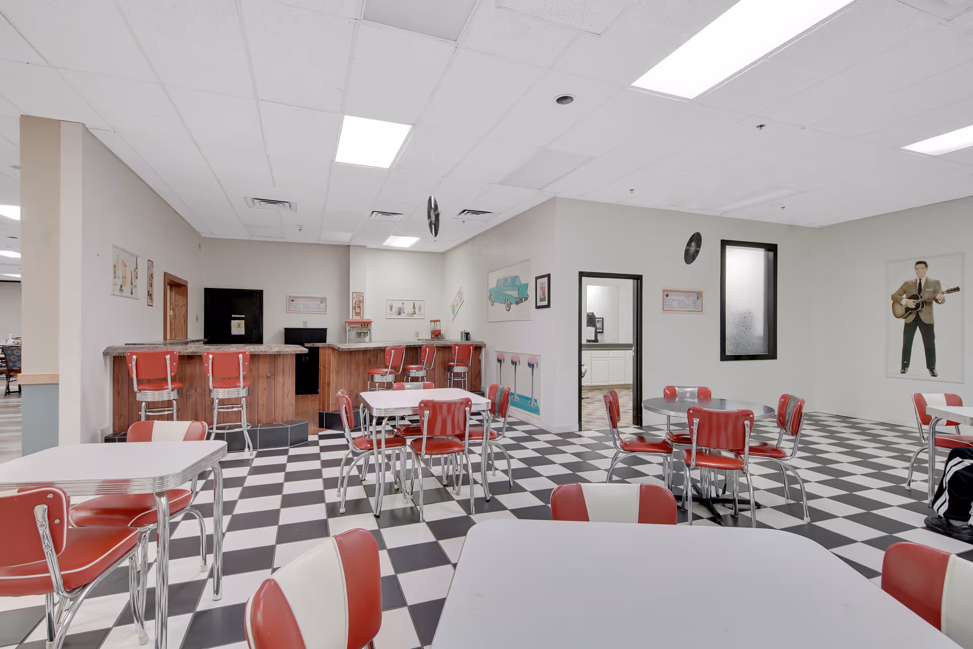 Retro diner-style common room with black-and-white checkered floor, red-and-white chairs and tables, and a wooden bar counter.