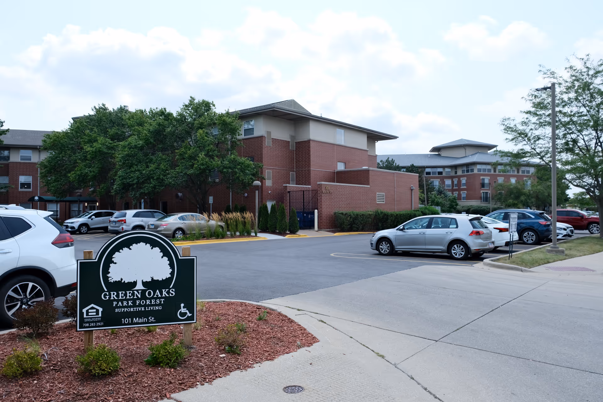 Exterior view of Green Oaks of Park Forest supportive living facility showing a parking lot with several cars, a green sign with the facility name and address, and multi-story brick buildings surrounded by trees and landscaping.