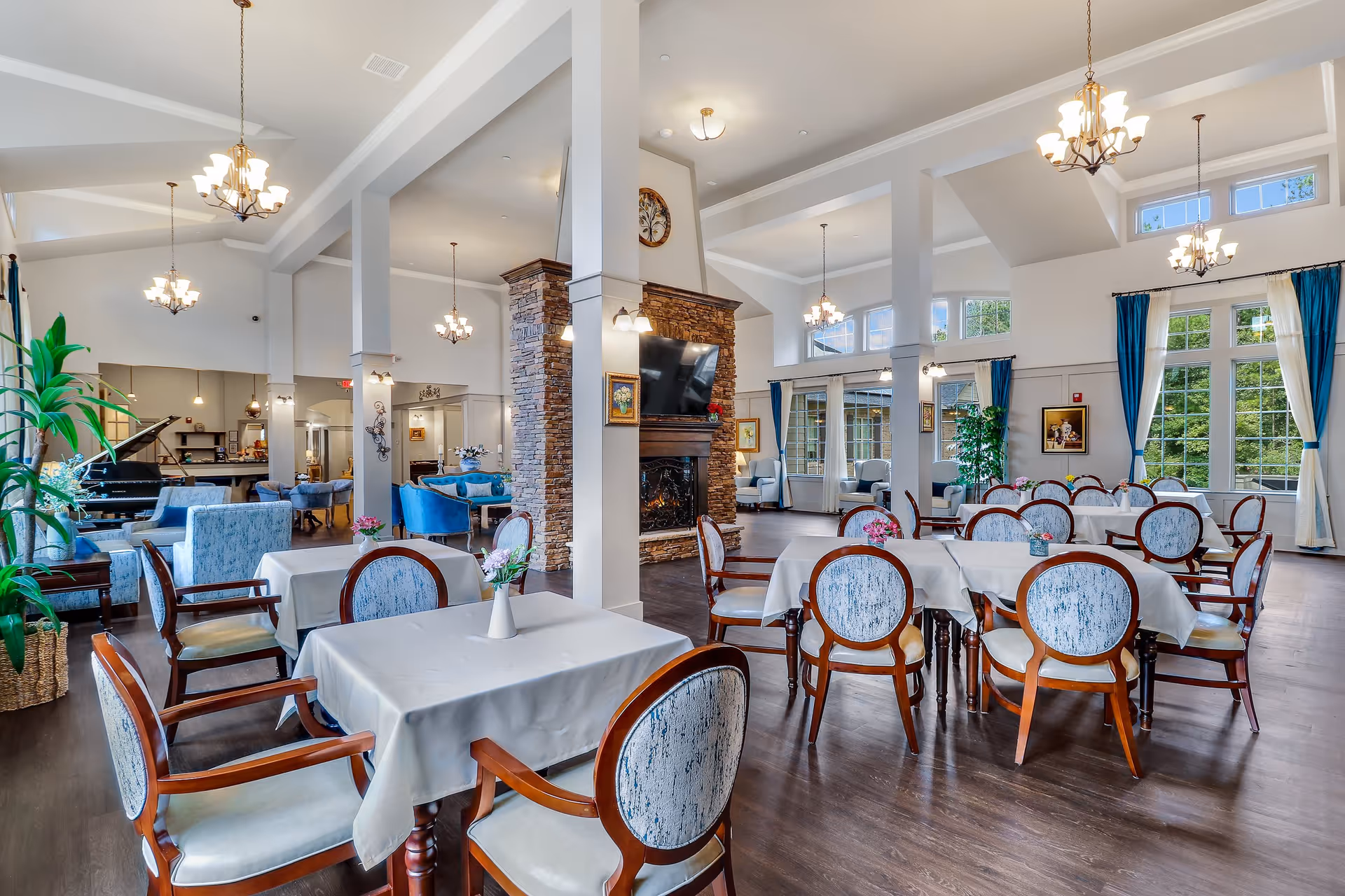 A spacious and elegant dining area in a senior living facility featuring multiple tables covered with white tablecloths and surrounded by wooden chairs with cushioned seats and backs. The room has high ceilings with several chandeliers, large windows with blue curtains allowing natural light, a stone fireplace with a mounted TV above it, and a seating area with blue upholstered chairs and a grand piano in the background.
