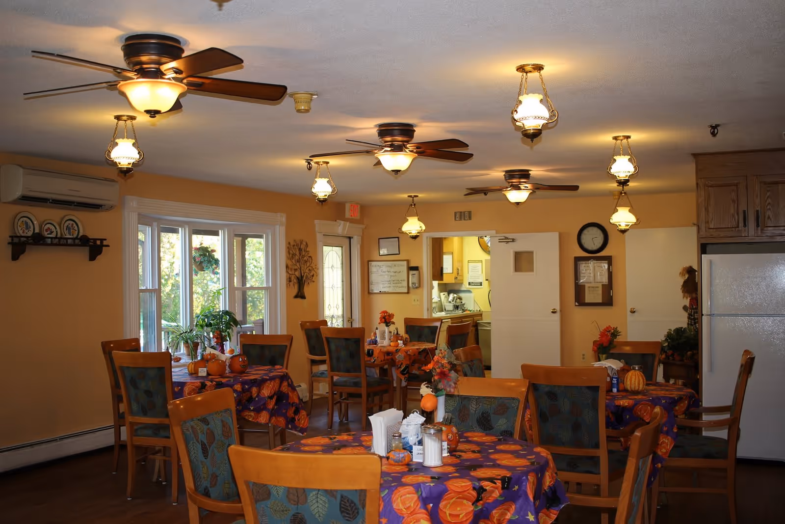 A cozy dining room in an assisted living facility with several round tables covered in festive tablecloths featuring pumpkins and autumn leaves. Each table has chairs with patterned upholstery, small decorative pumpkins, and floral centerpieces. The room has warm yellow walls, ceiling fans with lights, hanging lantern-style lights, a large window with plants on the sill, and a white refrigerator in the corner. A doorway leads to a kitchen area in the background.