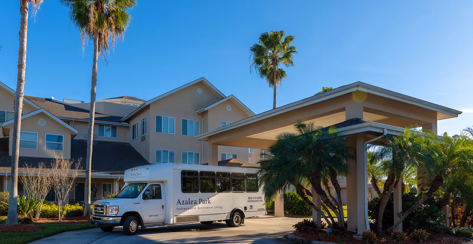 Exterior view of Azalea Park, a senior living community building with beige walls and multiple windows. A white shuttle bus with Azalea Park branding is parked under a covered entrance. Palm trees and landscaped greenery surround the area under a clear blue sky.
