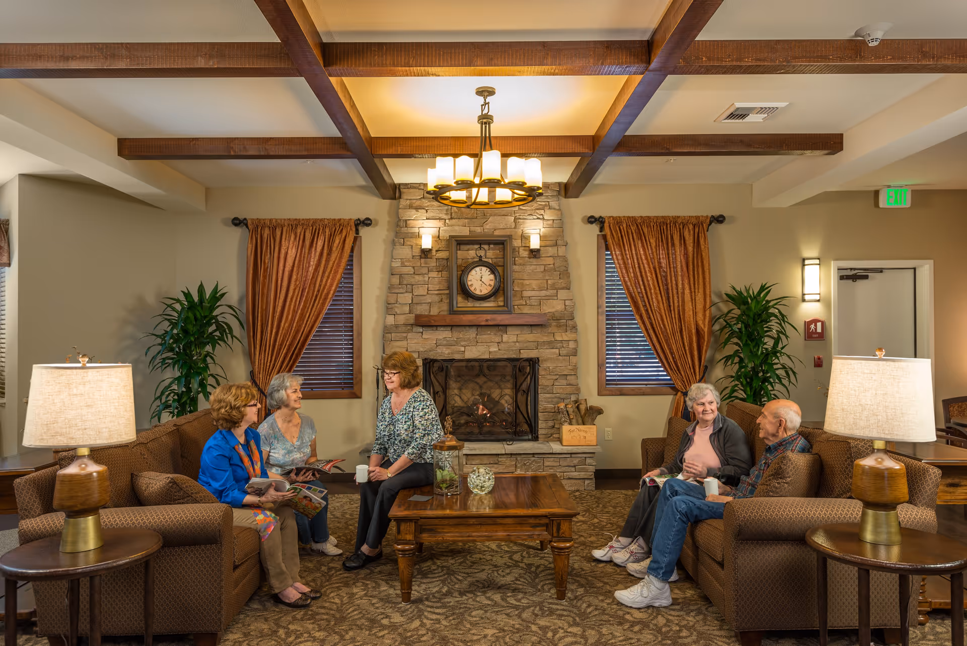 A cozy senior living facility common area with a stone fireplace in the center, flanked by two windows with brown curtains. Five elderly people are seated on two brown sofas facing each other, engaged in conversation and holding magazines and cups. The room features wooden ceiling beams, two table lamps on side tables, and decorative plants.