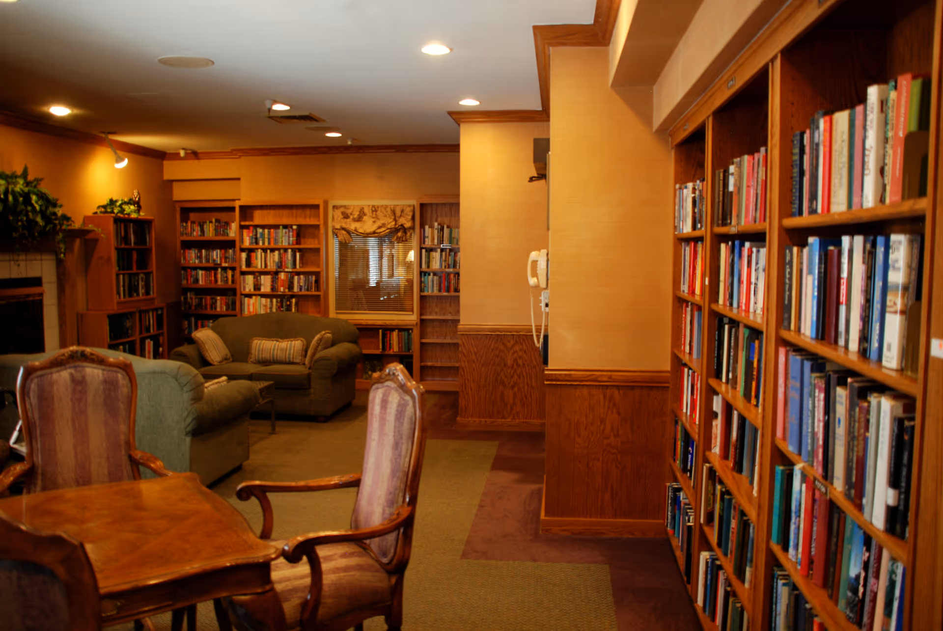 Cozy library room with wooden bookshelves filled with books, two upholstered armchairs, a wooden table with two chairs, and a green sofa with pillows. Warm lighting and wood paneling create a comfortable atmosphere.