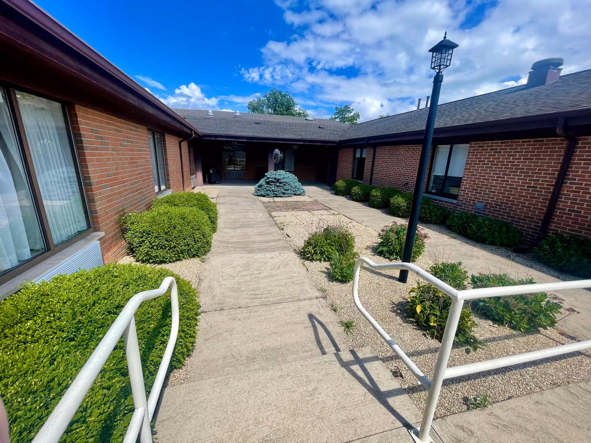 Outdoor courtyard area of a brick building with a concrete walkway, green bushes, small plants, and a black lamp post under a partly cloudy blue sky.
