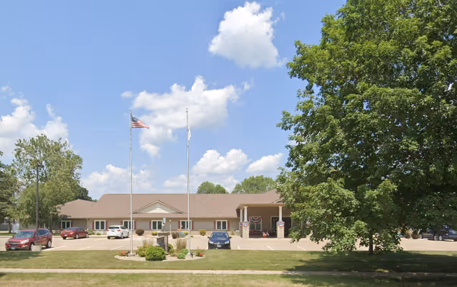 Front exterior view of Courtyard Estates of Knoxville, a single-story building with a pitched roof, two flagpoles with flags, a parking lot with several cars, and a large tree on the right side under a partly cloudy blue sky.